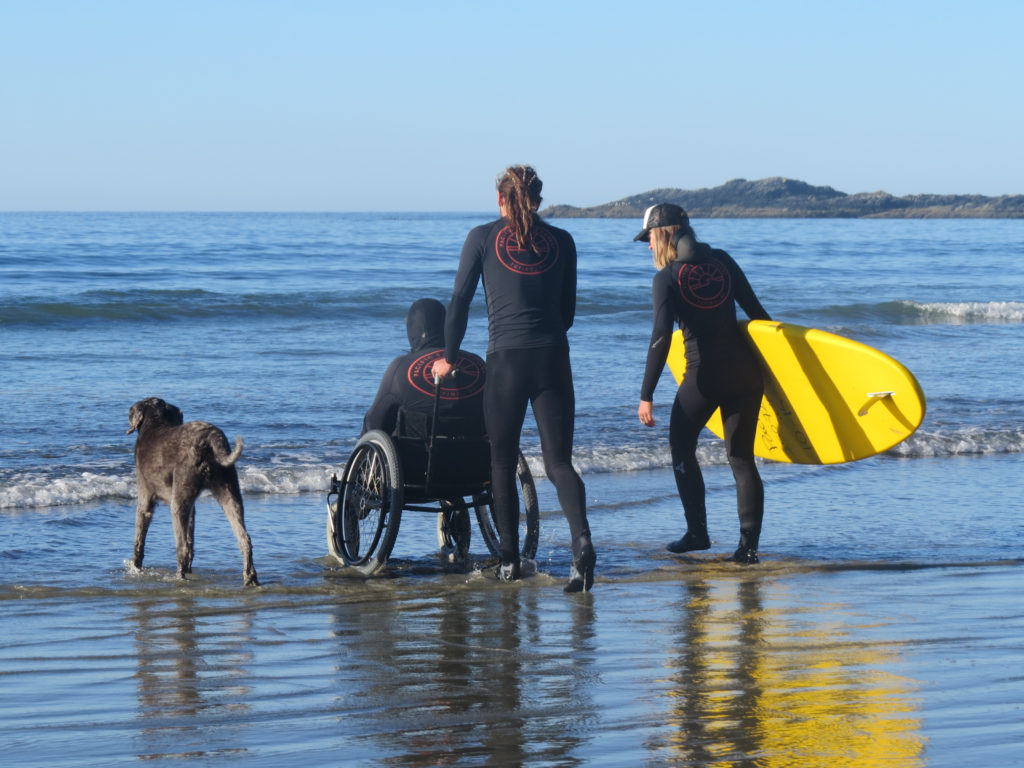 Two sufers wheeling a person in a wheelchair across the beach towards the ocean